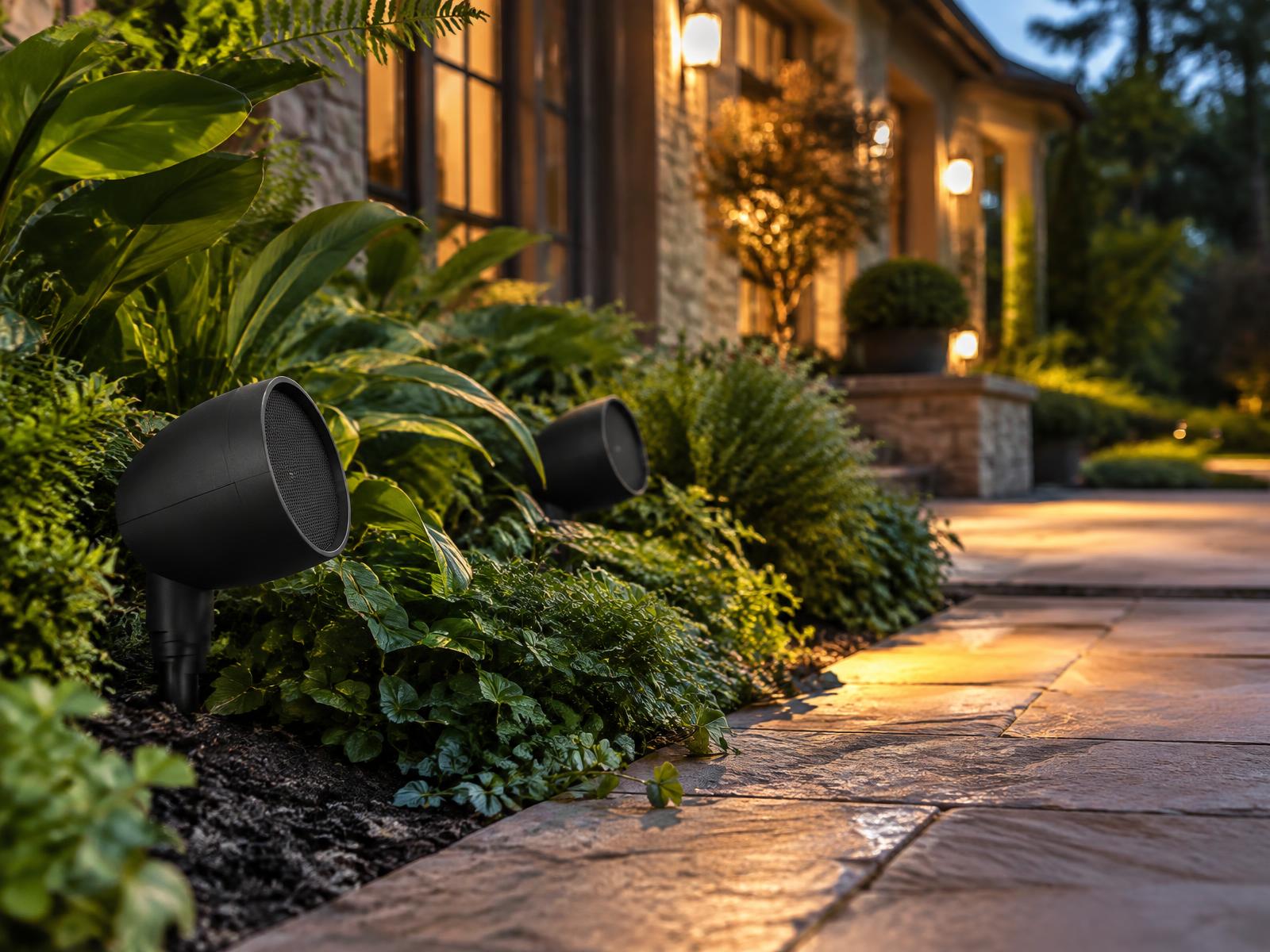 Black landscape speakers set into a planting bed beside a lit stone walkway at dusk